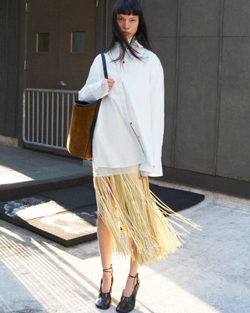 Cropped image of model standing wearing the Addy Shirt in Compact Cotton Poplin, the Veda Skirt in Faux Leather Fringe in Pale Yellow Multi, and the Glove Hinge Pumps in Black holding the Brant Bucket Bag in Soft Suede