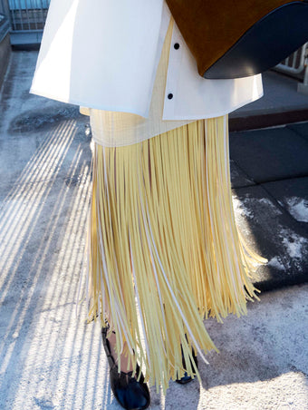 Cropped image of model standing wearing  the Addy Shirt in Compact Cotton Poplin, Veda Skirt in Faux Leather Fringe in Pale Yellow Multi, and the Glove Hinge Pumps in Black holding the Brant Bucket Bag in Soft Suede