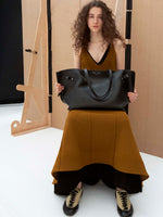 Image of model sitting wearing the Tavia Dress in Viscose Fringe Knit in Olive and the Arcadia Sneakers in Almond holding the Days Carryall in Smooth Calf in Black 