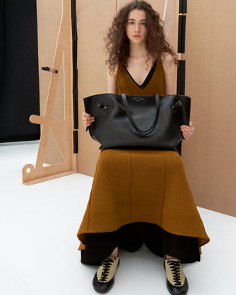 Cropped image of model sitting holding the a bag wearing the Tavia Dress in Viscose Fringe Knit in Olive and the Arcadia Sneakers in Almond holding the Days Carryall in Smooth Calf in Black 