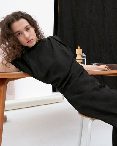 Cropped image of model laying against a table in the Hallie Dress in Hammered Viscose in Black