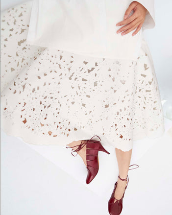 Cropped image of model laying down wearing the Olene Skirt in Broderie Anglaise in White and the Glove Hinge Pumps in Merlot