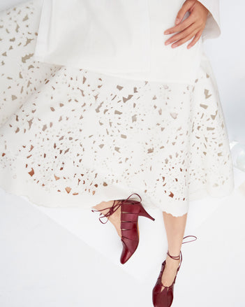 Cropped image of model laying down wearing the Olene Skirt in Broderie Anglaise in White and the Glove Hinge Pumps in Merlot