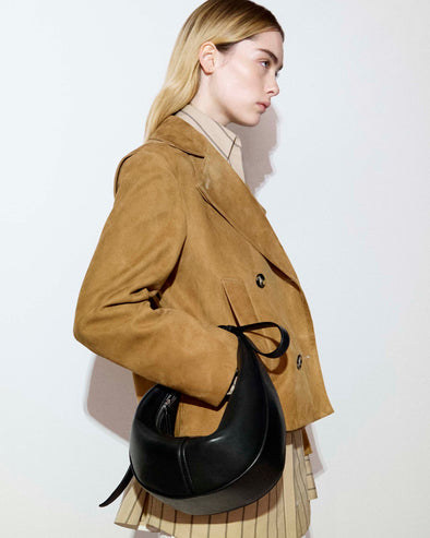 Cropped image of the side of a model standing against a white backdrop in the Addy Top in Stripe Cotton Silk in Taupe, Nola Jacket in Suede in Pony, carrying the Mini Slide Bag in Black