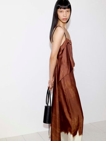 Cropped model standing against a white wall wearing the Wilma Dress in Double Printed Silk Twill in Sienna Multi holding the Cross Body Silo in Black