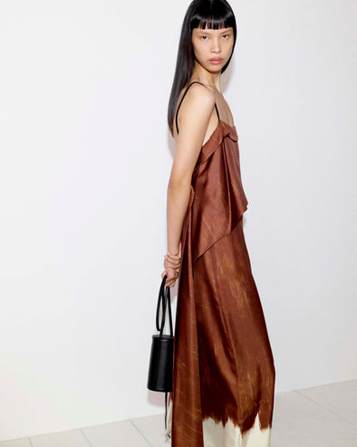 Cropped model standing against a white wall wearing the Wilma Dress in Double Printed Silk Twill in Sienna Multi holding the Cross Body Silo in Black
