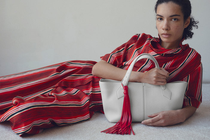 Image of model Elise Kouzou wearing red striped set holding the white East West tote laying on a white carpet against a white wall 