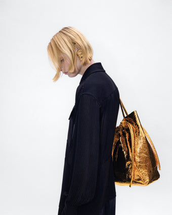 4x5 image of a model's left hand profile, wearing a gold drawstring tote over her shoulder with a black shirtdress against a white background 