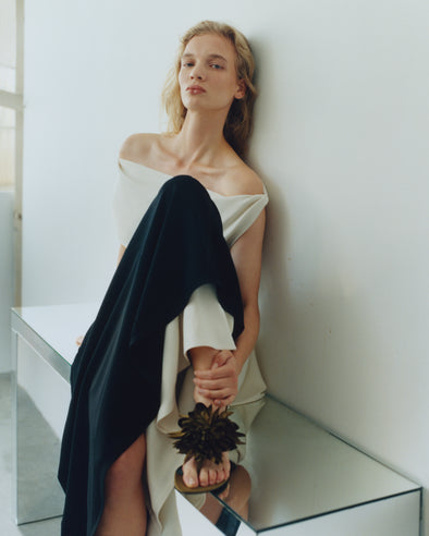 Proenza Schouler image of model sitting on mirrored table wearing the Daliah Dress in Satin Back Crepe in Black/Ecru with the Tee Thong Flower Sandals in Satin in Pstachio