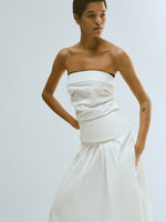 Cropped editorial image of model standing in front of blank white wall wearing the Maisy Strapless Top in Poplin in White with the Nessa Skirt in Poplin in White