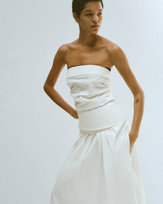 Cropped editorial image of model standing in front of blank white wall wearing the Maisy Strapless Top in Poplin in White with the Nessa Skirt in Poplin in White