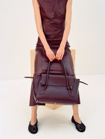 Cropped image of model wearing Backless Leather Dress in Ox Blood with Tee Perforated Ballerina Flats in black, carrying Tate Bag in dark red 
