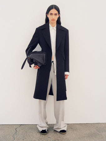 Image of model wearing Thea Coat in Wool Gabardine Suiting in black and Savannah Pant in Wool Gabardine Suiting in taupe with Track Sneakers in black/white, carrying Tate Bag in black