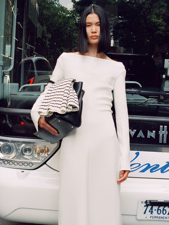 Cropped image of model leaning against bus, wearing Yara Dress In Bi-Stretch Suiting in off white, carrying Tate bag in black