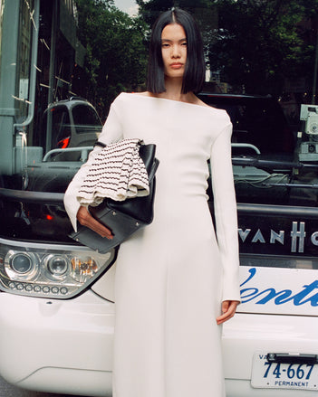Cropped image of model leaning against bus, wearing Yara Dress In Bi-Stretch Suiting in off white, carrying Tate bag in black