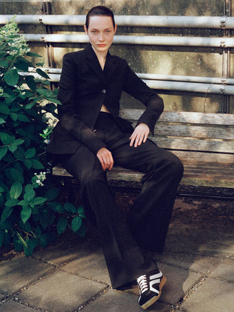 Image of model sitting on park bench wearing Lark Blazer and Savannah Pant in Wool Gabardine Suiting in black with Track Sneakers in black