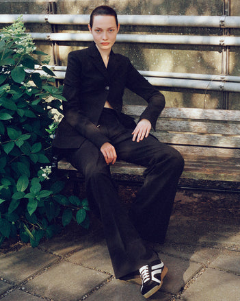 Image of model sitting on park bench wearing Lark Blazer and Savannah Pant in Wool Gabardine Suiting in black with Track Sneakers in black