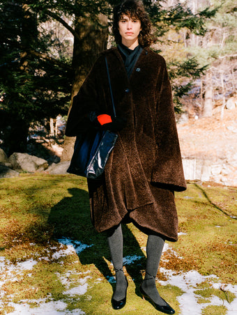Image of model standing in snowy field of moss wearing Varretta Coat in Chocolate Alpaca Coating, Ysabelle Top in Anthracite Silk Viscose, Argyle Gloves in Grey Cashmere, Reade Shoulder Bag in Black Paper Calf, and Uma Tie Pumps in Black Slick Calf