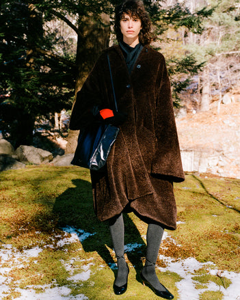 Image of model standing in snowy field of moss wearing Varretta Coat in Chocolate Alpaca Coating, Ysabelle Top in Anthracite Silk Viscose, Argyle Gloves in Grey Cashmere, Reade Shoulder Bag in Black Paper Calf, and Uma Tie Pumps in Black Slick Calf
