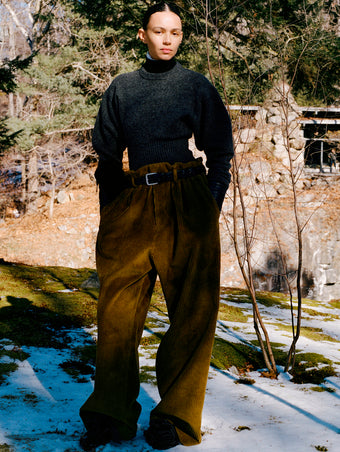 Model standing in snowy field of moss wearing Randi Sweater in Dark Grey Boiled Wool, Fox Sweater in Black Viscose Rib, Salome Pant in Fatigue Corduroy, Silo Clutch in Chocolate Shearling, Braid Belt in Black Calf, and Tee Souvenir Sandals in Black Satin