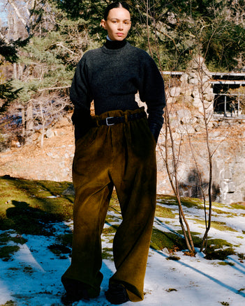 Model standing in snowy field of moss wearing Randi Sweater in Dark Grey Boiled Wool, Fox Sweater in Black Viscose Rib, Salome Pant in Fatigue Corduroy, Silo Clutch in Chocolate Shearling, Braid Belt in Black Calf, and Tee Souvenir Sandals in Black Satin