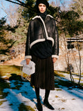 Model standing in snowy field wearing Alya Jacket in Black/Off White Shearling, Solene Top in Anthracite Silk Viscose, Eda Skirt in Espresso Wool Jersey, Beret in Black Wool Knit, Reade Shoulder Bag in Optic White Paper Calf, and Uma Tie Pumps in Black Slick Calf