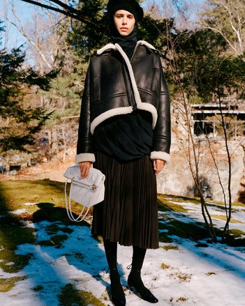 Model standing in snowy field wearing Alya Jacket in Black/Off White Shearling, Solene Top in Anthracite Silk Viscose, Eda Skirt in Espresso Wool Jersey, Beret in Black Wool Knit, Reade Shoulder Bag in Optic White Paper Calf, and Uma Tie Pumps in Black Slick Calf