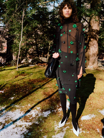 Model standing on snowy field of moss wearing Lex Top in Black/Green Embroidered Viscose Georgette, Lex Skirt in Black/Green Embroidered Viscose Georgette, Reade Shoulder Bag in Black Paper Calf, and Slash Slingbacks in Optic White Spazzolato