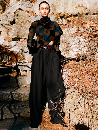 Model standing against rock staircase and foliage wearing Vanne Top in Brown Argyle Velvet Knit, Salome Pant in Black Classic Twill, Silo Clutch in Black Shiny Calf, Braid Belt in Bordeaux Croc Embossed Calf, and Uma Ankle Boots in Black Slick Calf