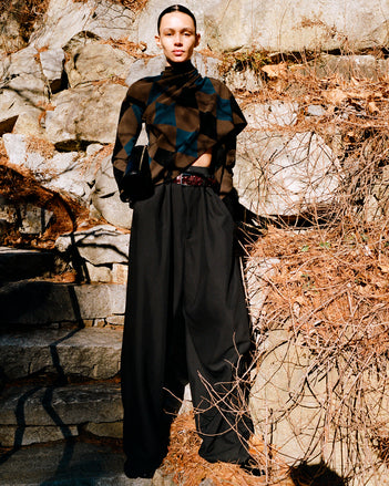 Model standing against rock staircase and foliage wearing Vanne Top in Brown Argyle Velvet Knit, Salome Pant in Black Classic Twill, Silo Clutch in Black Shiny Calf, Braid Belt in Bordeaux Croc Embossed Calf, and Uma Ankle Boots in Black Slick Calf