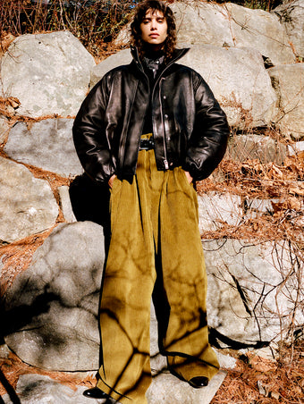 Model standing against stone wall and foliage wearing Laney Jacket in Black Semigloss Leather, Jeanne Sweater in Black Eco Cashmere, Lynsey Top in Black/White Viscose Striped Shirting, Fox Sweater in Black Viscose Rib, Salome Pant in Fatigue Corduroy, Braid Belt in Black Calf, and Uma Ankle Boots in Black Slick Calf