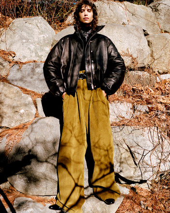 Model standing against stone wall and foliage wearing Laney Jacket in Black Semigloss Leather, Jeanne Sweater in Black Eco Cashmere, Lynsey Top in Black/White Viscose Striped Shirting, Fox Sweater in Black Viscose Rib, Salome Pant in Fatigue Corduroy, Braid Belt in Black Calf, and Uma Ankle Boots in Black Slick Calf