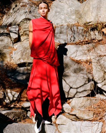 Model standing against rock wall and foliage wearing Deise Dress in Red Viscose Crepe and Slash Slingbacks in Optic White Spazzolato