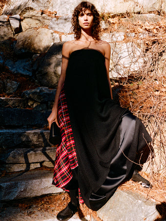 Model standing against stone staircase and wall wearing Elea Dress in Rust Boucle Plaid Suiting, Silo Clutch in Black Shiny Calf, and Arcadia Sneakers in Black Calfsuede