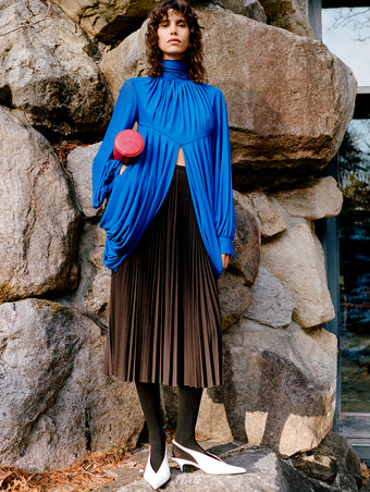Model standing against stone wall wearing Bari Top in Cobalt Gauzy Jersey, Eda Skirt in Espresso Wool Jersey, Silo Clutch in Red Shiny Calf, and Slash Slingbacks in Optic White Spazzolato