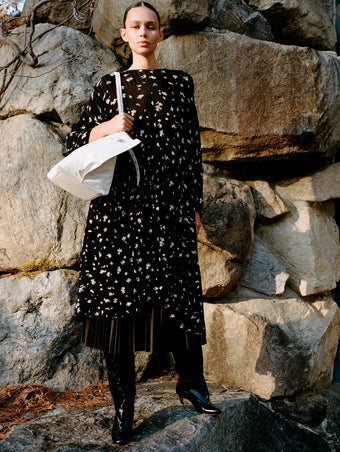 Model standing against stone wall wearing Zeda Dress in Black/White Acid Print Knit, Eda Skirt in Espresso Wool Jersey, Days Carryall in Optic White Paper Calf, and Uma Over the Knee Boots in Black Slick Calf