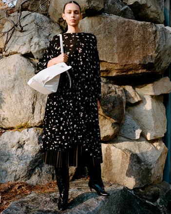 Model standing against stone wall wearing Zeda Dress in Black/White Acid Print Knit, Eda Skirt in Espresso Wool Jersey, Days Carryall in Optic White Paper Calf, and Uma Over the Knee Boots in Black Slick Calf