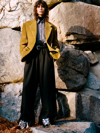 Model standing against rock wall and foliage wearing Daria Jacket in Fatigue Corduroy, Lynsey Top in Black/White Viscose Striped Shirting, Fox Sweater in Black Viscose Rib, Salome Pant in Black Classic Twill, Braid Belt in Black Calf, and Tee Souvenir Sandals in Silver Metallic Foil