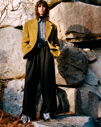 Model standing against rock wall and foliage wearing Daria Jacket in Fatigue Corduroy, Lynsey Top in Black/White Viscose Striped Shirting, Fox Sweater in Black Viscose Rib, Salome Pant in Black Classic Twill, Braid Belt in Black Calf, and Tee Souvenir Sandals in Silver Metallic Foil