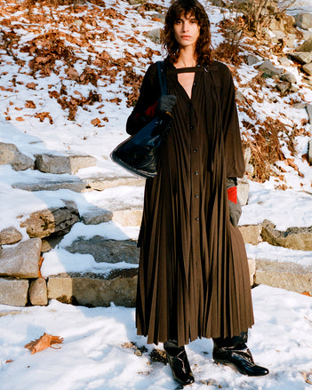 Model standing on snowy stone steps wearing Fanni Dress in Espresso Wool Jersey, Argyle Gloves in Grey Cashmere, Days Carryall in Black Paper Calf, and Uma Over the Knee Boots in Black Slick Calf