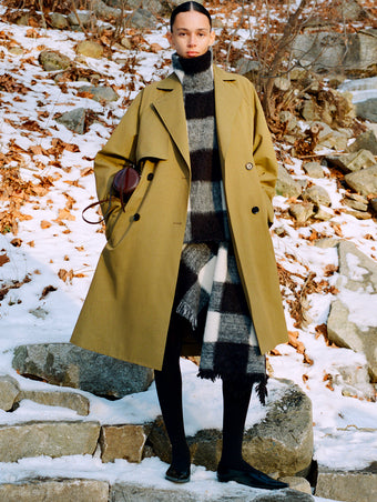 Model standing on snowy stone steps wearing Adelaide Coat in Moss Cotton Viscose, Kai Top in Black Brushed Glenplaid Knit, Kai Skirt in Black Brushed Glenplaid Knit, Crossbody Silo in Cedar Shiny Calf, and Slash Flats in Black Spazzolato