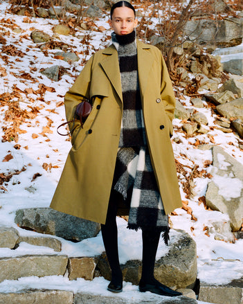 Model standing on snowy stone steps wearing Adelaide Coat in Moss Cotton Viscose, Kai Top in Black Brushed Glenplaid Knit, Kai Skirt in Black Brushed Glenplaid Knit, Crossbody Silo in Cedar Shiny Calf, and Slash Flats in Black Spazzolato