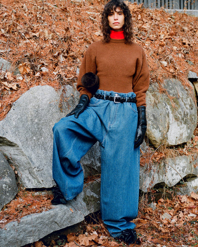 Model standing with one foot up on rock siding wearing Randi Sweater in Brown Boiled Wool, Fox Sweater in Red Viscose, Salome Jean in Medium Blue Lightweight Denim, Silo Clutch in Chocolate Shearling, and Tee Souvenir Sandals in Black Satin