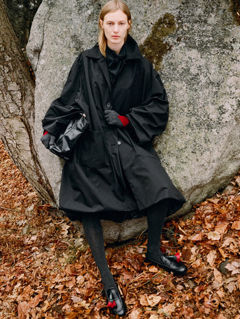 Model leaning against boulder standing in falling leaves wearing Verretta Coat in Black Coated Cotton, Ysabelle Top in Anthracite Silk Viscose, Argyle Gloves in Grey Cashmere, Reade Shoulder Bag in Black Paper Calf, and Albers Loafer in Black Box Calf