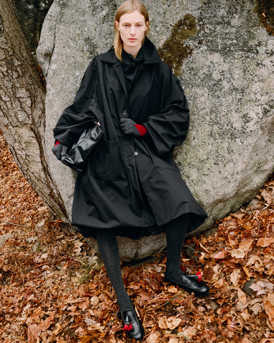 Model leaning against boulder standing in falling leaves wearing Verretta Coat in Black Coated Cotton, Ysabelle Top in Anthracite Silk Viscose, Argyle Gloves in Grey Cashmere, Reade Shoulder Bag in Black Paper Calf, and Albers Loafer in Black Box Calf