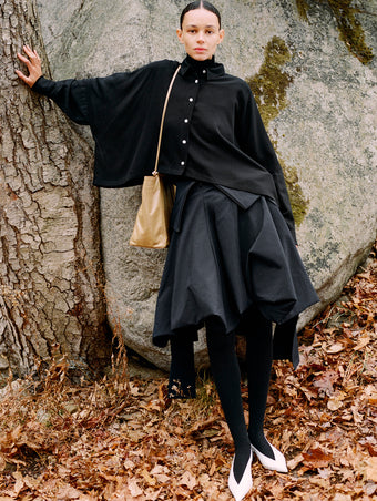 Model leaning against stone boulder wearing Tiana Dress in Black Technical Canvas, Fox Sweater in Black Viscose Rib, Zad Top in Black Classic Twill, Reade Shoulder Bag in Tan Paper Calf, and Slash Slingbacks in Optic White Spazzolato