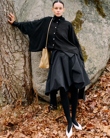 Model leaning against stone boulder wearing Tiana Dress in Black Technical Canvas, Fox Sweater in Black Viscose Rib, Zad Top in Black Classic Twill, Reade Shoulder Bag in Tan Paper Calf, and Slash Slingbacks in Optic White Spazzolato