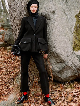 Model standing against stone boulder in fallen leaves wearing Silvia Jacket in Black Wool Scuba, Ismene Pant in Black Wool Scuba, Solene Top in Anthracite Silk Viscose, Beret in Black Wool Knit, Reade Shoulder Bag in Black Paper Calf, and Albers Loafer in Black Box Calf