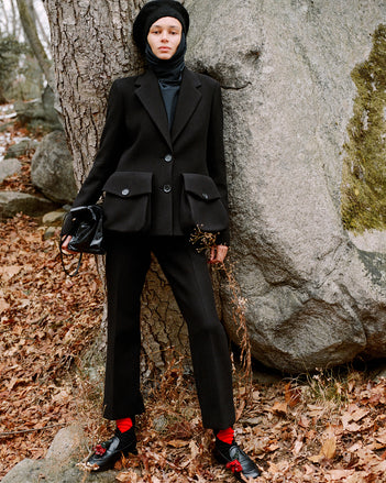 Model standing against stone boulder in fallen leaves wearing Silvia Jacket in Black Wool Scuba, Ismene Pant in Black Wool Scuba, Solene Top in Anthracite Silk Viscose, Beret in Black Wool Knit, Reade Shoulder Bag in Black Paper Calf, and Albers Loafer in Black Box Calf