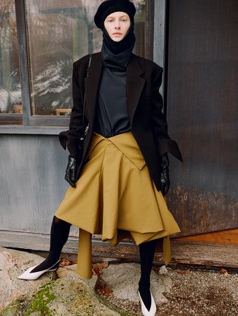 Model standing against the side of a window wearing Daria Jacket in Black Wool Scuba, Solene Top in Anthracite Silk Viscose, Tiana Dress in Fatigue Technical Canvas, Beret in Black Wool Knit, Reade Shoulder Bag in Black Paper Calf, and Slash Slingbacks in Optic White Spazzolato